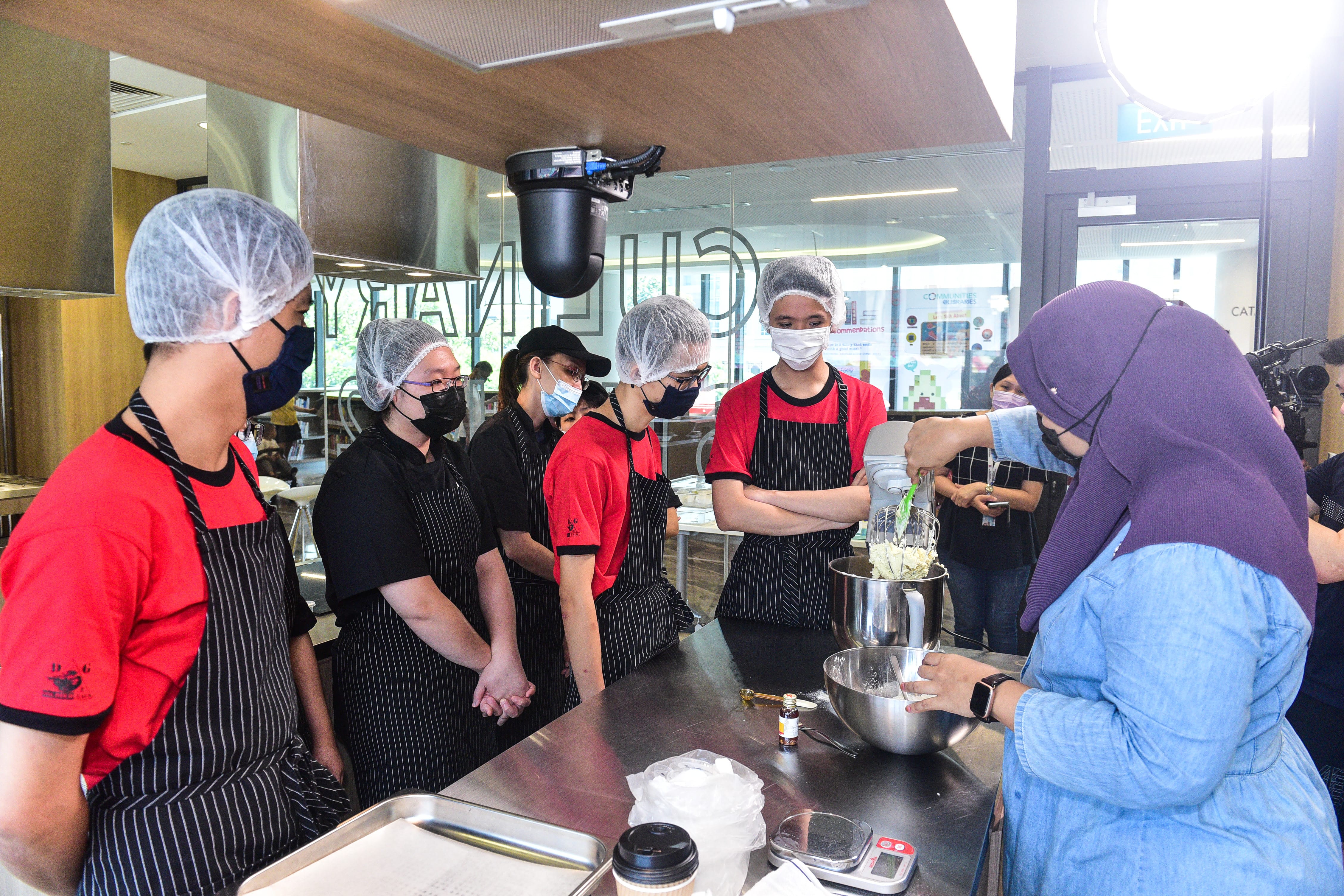 A group of individuals gathered around a kitchen island, watching a baker demonstrate baking techniques.